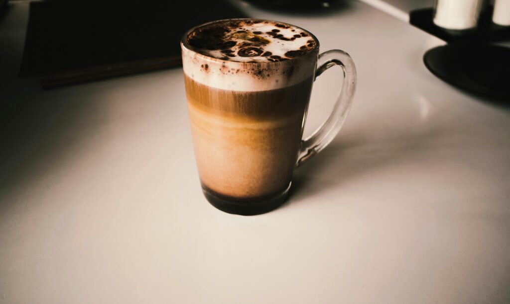 Warm cappuccino with frothy cream and chocolate sprinkles in a glass mug on a minimalist table.